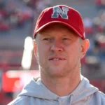 Scott Frost wearing a red Nebraska cap and gray hoodie, standing on the football field during a game.