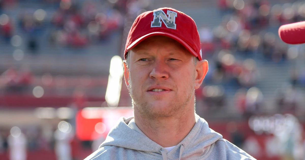 Scott Frost wearing a red Nebraska cap and gray hoodie, standing on the football field during a game.