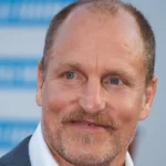 Woody Harrelson smiling in suit with blue background - actor with $70 million net worth